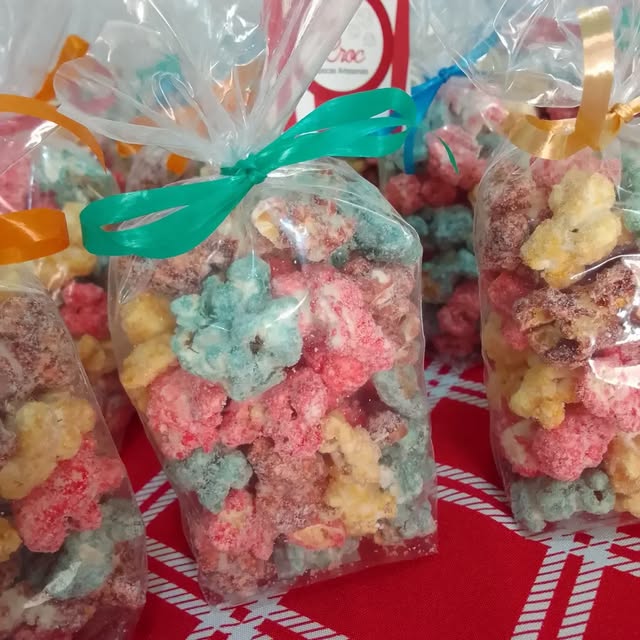 Sacos de pipoca colorida com laços verde, laranja e dourado, sobre toalha vermelha.